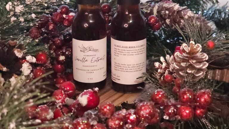 Two bottles of homemade vanilla extract surrounded by Christmas decorations including pine branches, red berries, and a pinecone